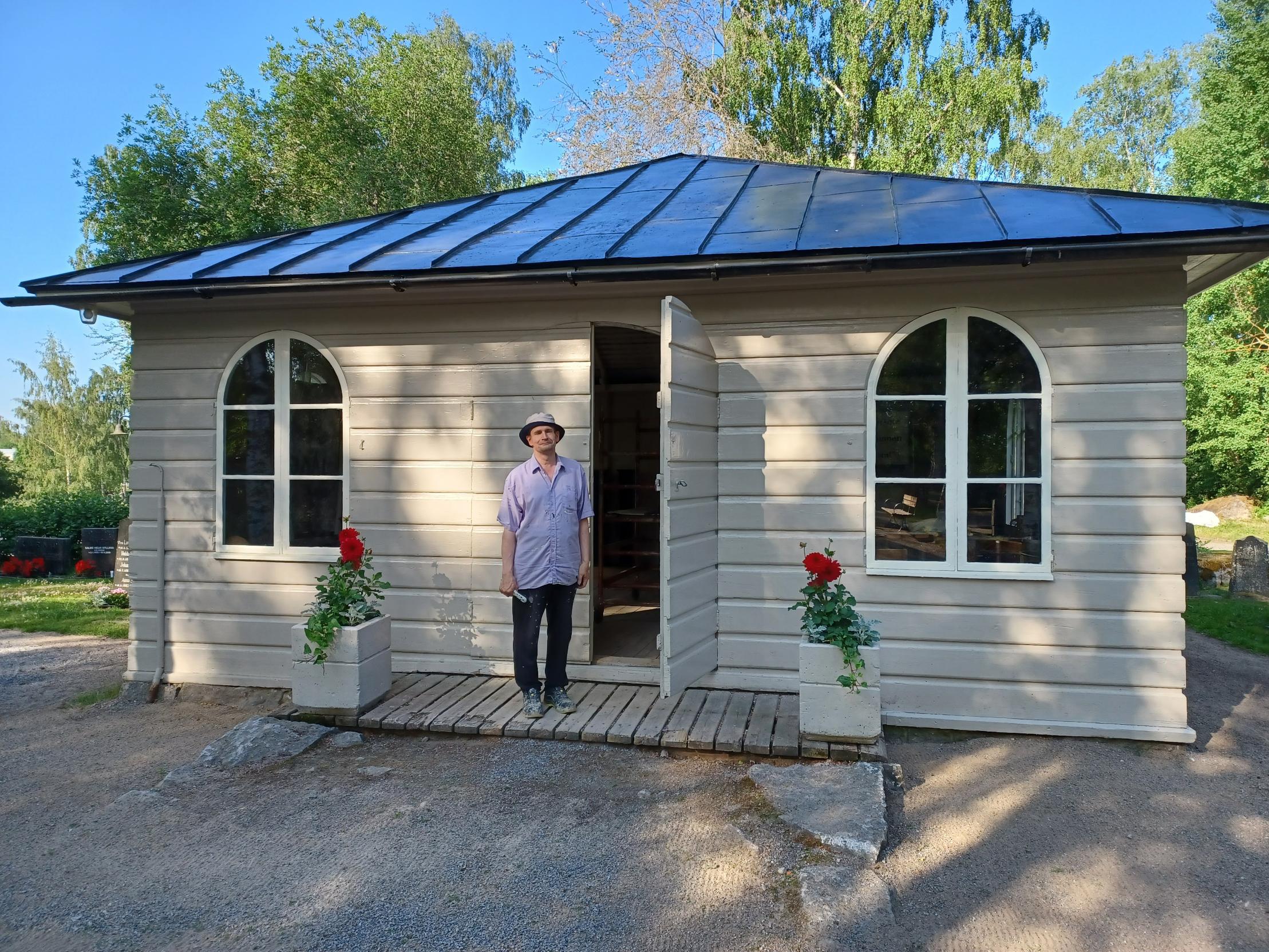 Man i ljus skjorta står framför en liten vacker gammal byggnad. Solen skiner och det är sommar.